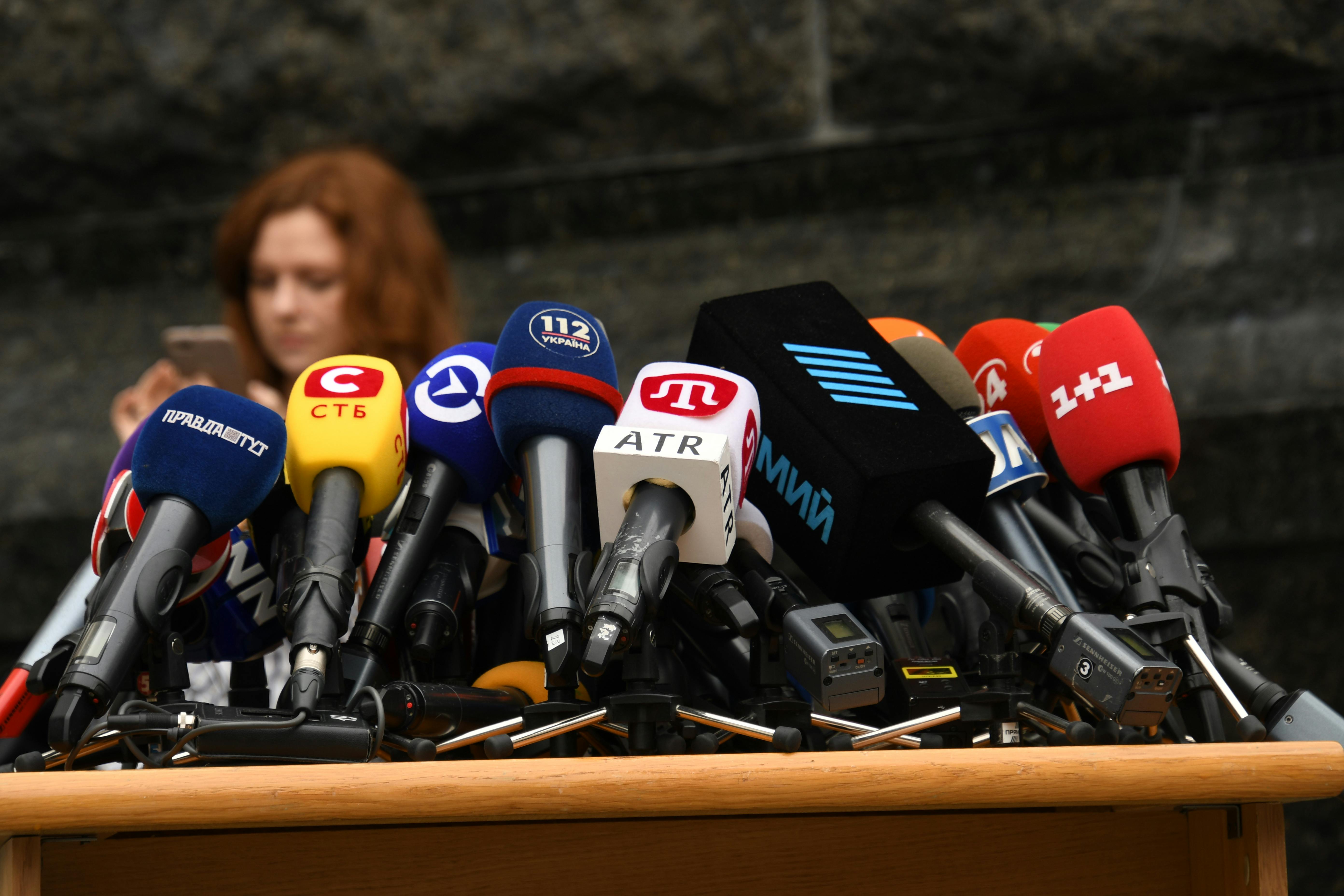 Multiple microphones from various news outlets arranged on a podium outdoors, suggesting a press conference or media event; the background is neutral and the atmosphere feels serious and anticipatory; visible microphone covers display logos and Cyrillic text including ATR, 112 Ukraine, and 1+1