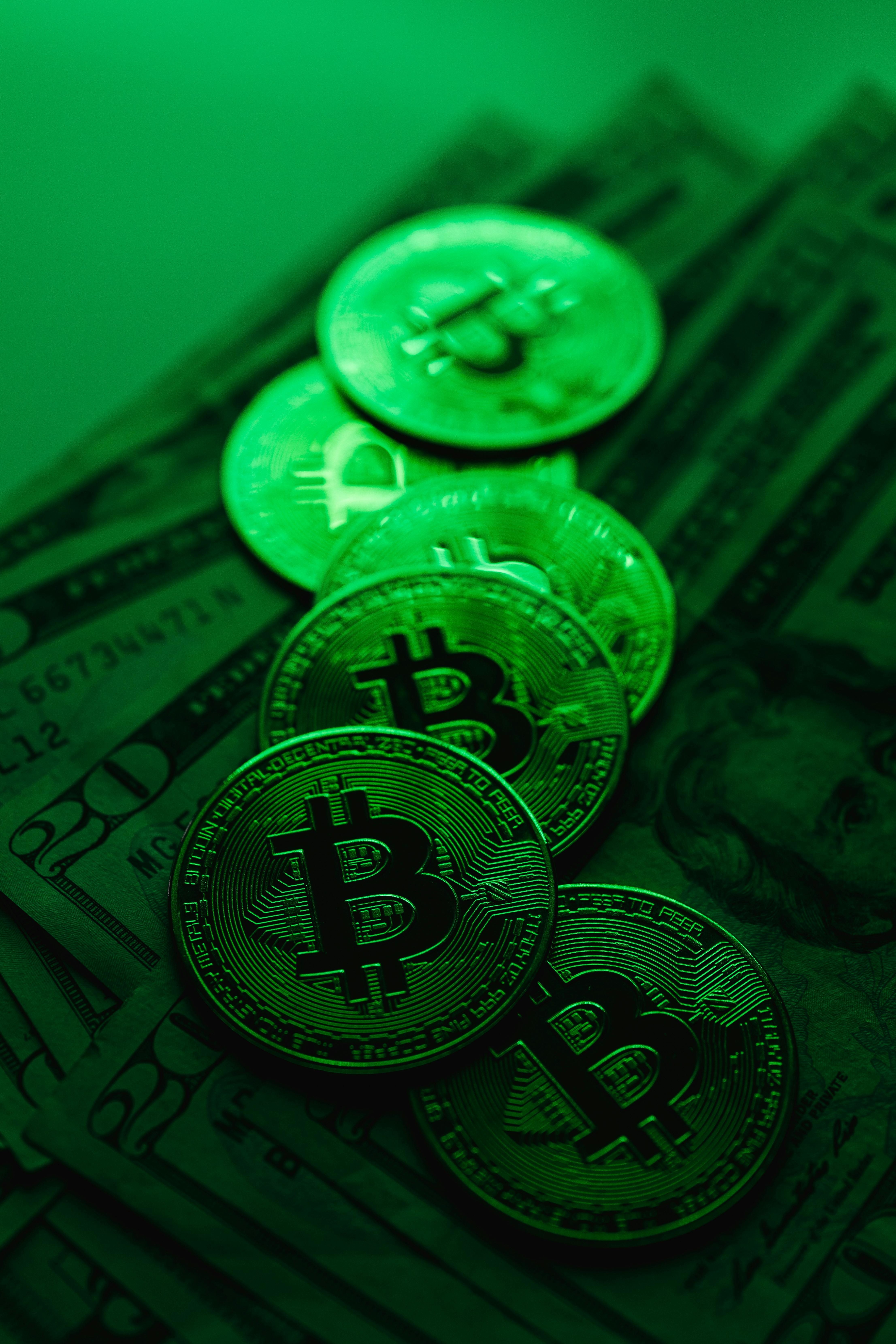 Several bitcoin coins placed on top of US dollar bills under a green light, highlighting the intersection of cryptocurrency and traditional finance; the scene feels futuristic and energetic