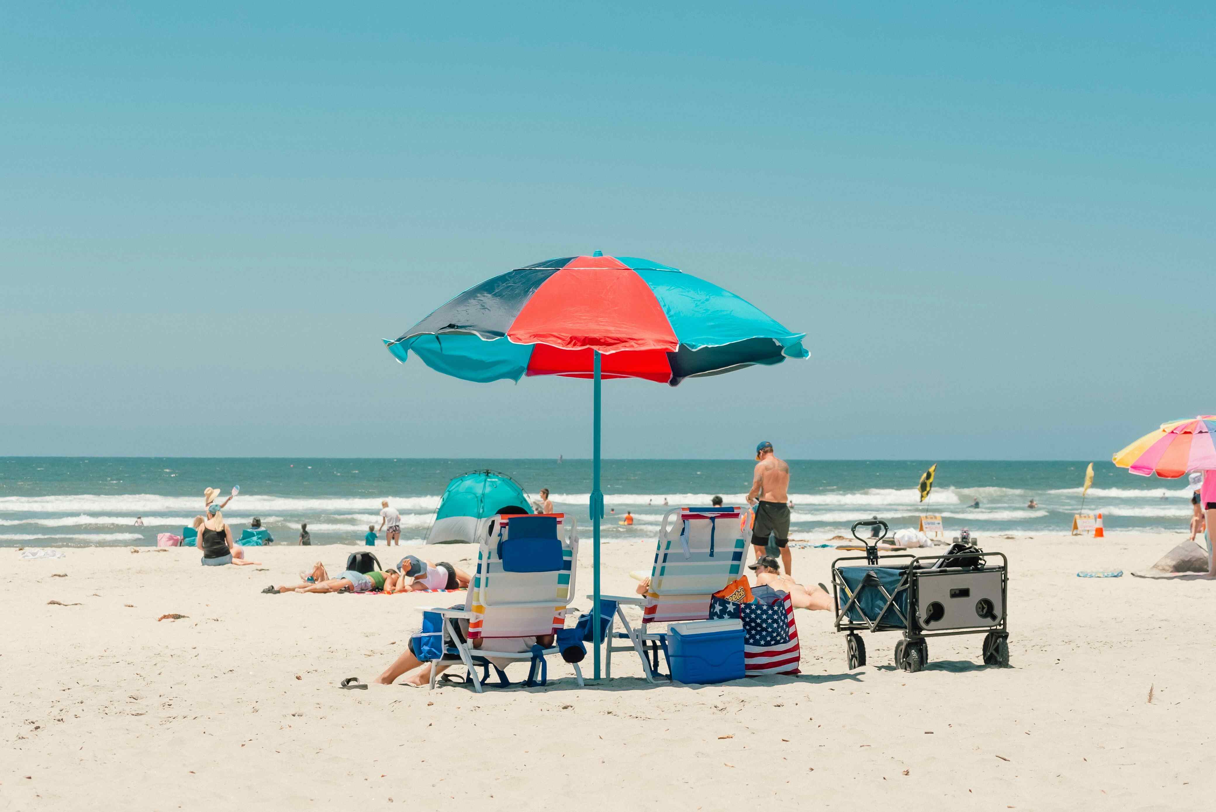 Two beach chairs and a colorful umbrella on a sunny sandy beach with people relaxing and playing near the ocean; the scene feels relaxed and joyful